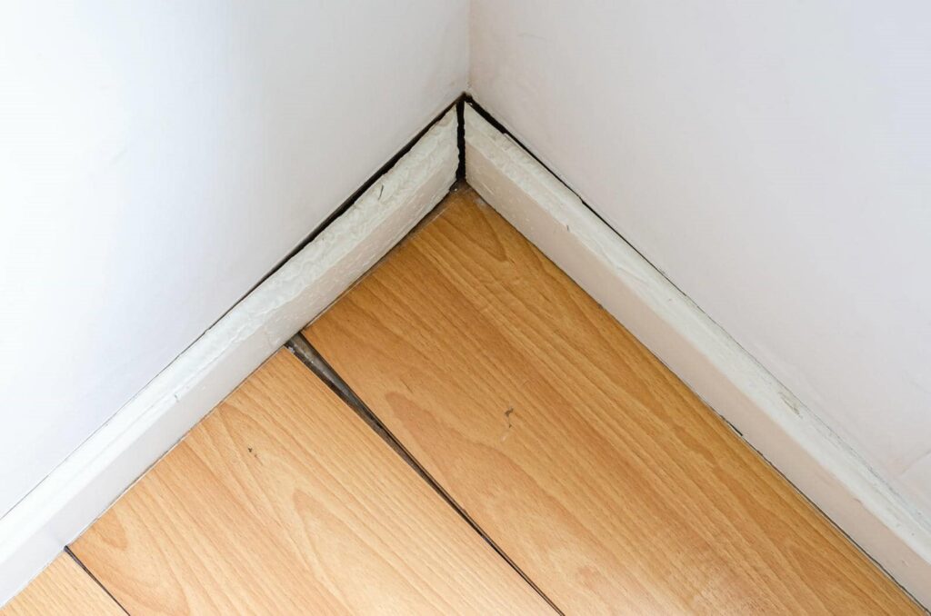 Corner of a room with wooden floorboards and white baseboards showing gaps and water damage