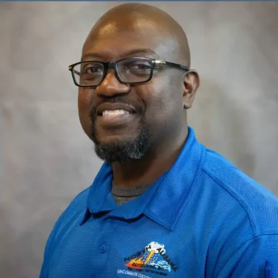 Man with glasses smiling, wearing a blue collared shirt against a gray background.
