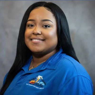 Smiling woman with long black hair wearing a blue polo shirt against a gray background.