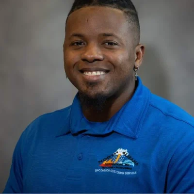 Smiling man wearing a blue polo shirt with a logo and text on the chest against a gray background.