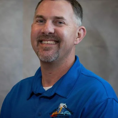 Smiling man with short hair and goatee wearing a blue collared shirt against a gray background.
