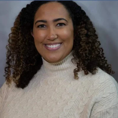 Smiling woman with curly hair wearing a cream cable-knit turtleneck sweater against a gray background.